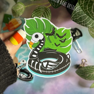 May include: Black and white skeleton flamingo pool float with a green tropical leaf and bats. The sticker says "Spookshow Babe Designs".