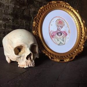 May include: A framed watercolour painting of a person with a flower crown and an eye in their chest. The painting is in a gold oval frame and is sitting on a dark wooden surface next to a human skull.