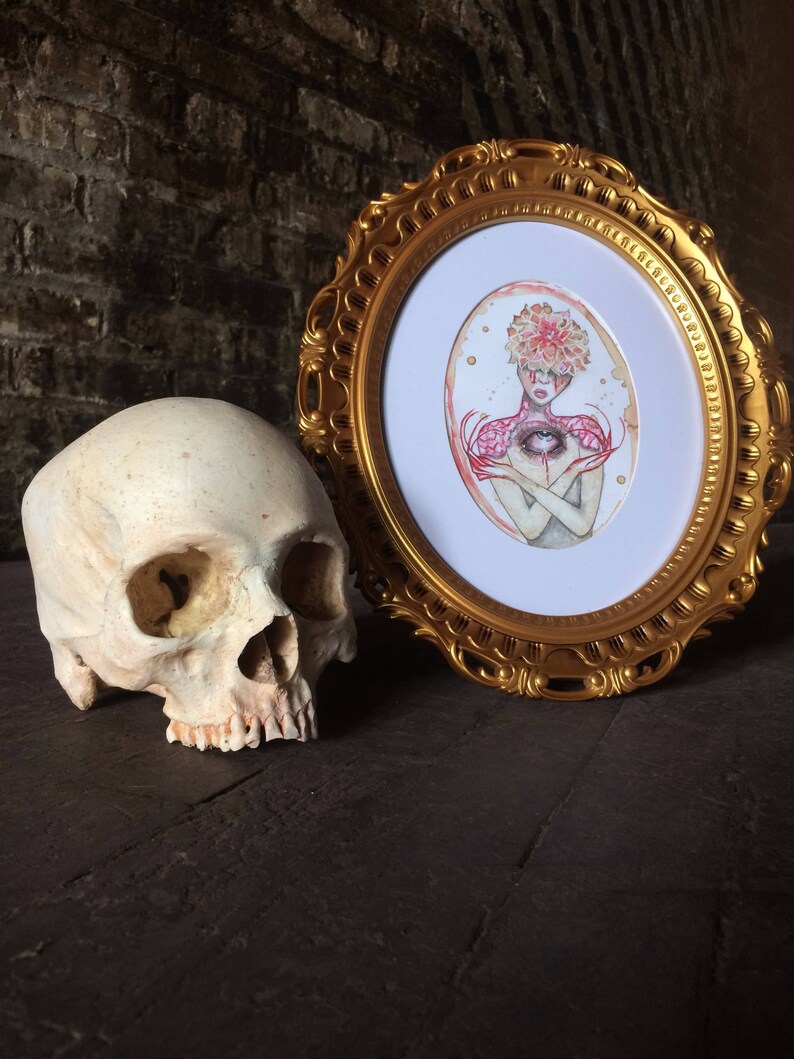 May include: A framed watercolour painting of a person with a flower crown and an eye in their chest. The painting is in a gold oval frame and is sitting on a dark wooden surface next to a human skull.