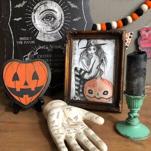 Peut inclure: Une nature morte sur le th&egrave;me d'Halloween avec une citrouille en bois, une illustration encadr&eacute;e d'une sorci&egrave;re, une main de diseuse de bonne aventure et une bougie noire sur un support turquoise. L'illustration repr&eacute;sente une sorci&egrave;re avec un chapeau noir et une citrouille. La main de diseuse de bonne aventure a des lignes et des symboles sur la paume.