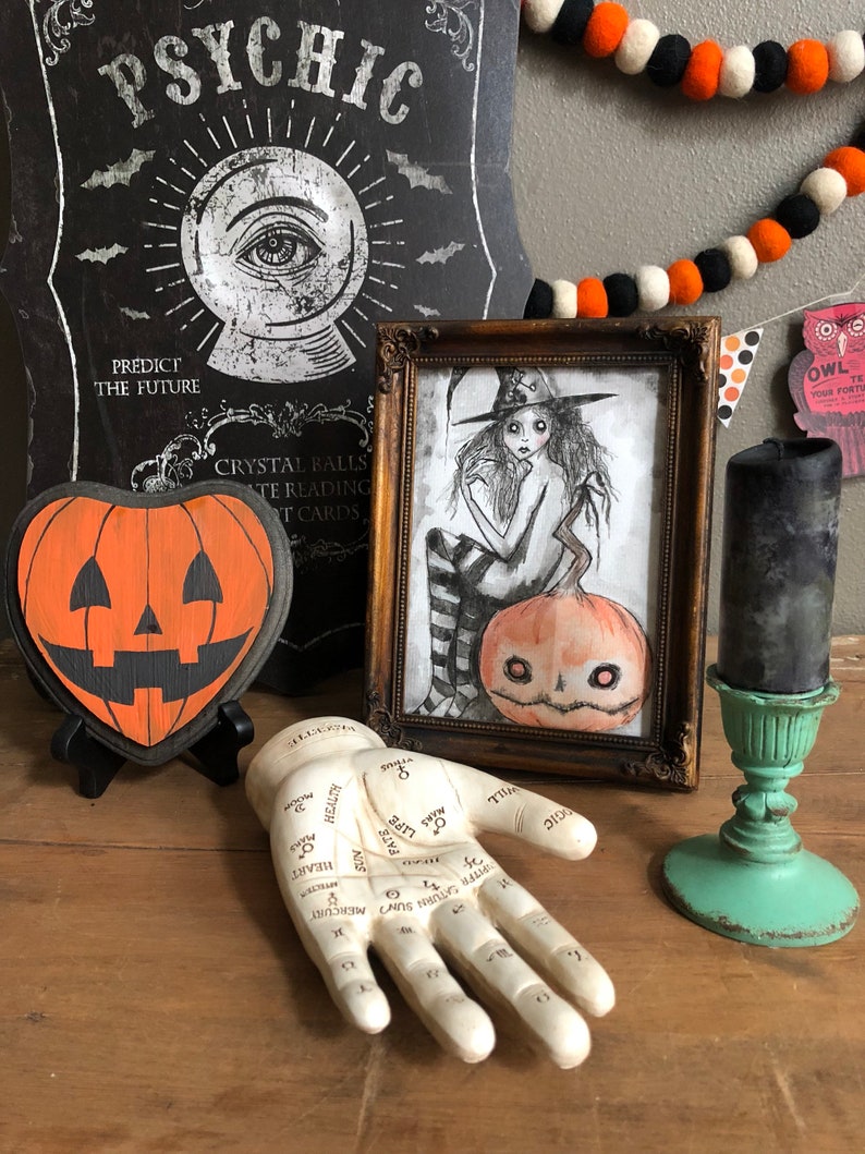 Peut inclure: Une nature morte sur le th&egrave;me d'Halloween avec une citrouille en bois, une illustration encadr&eacute;e d'une sorci&egrave;re, une main de diseuse de bonne aventure et une bougie noire sur un support turquoise. L'illustration repr&eacute;sente une sorci&egrave;re avec un chapeau noir et une citrouille. La main de diseuse de bonne aventure a des lignes et des symboles sur la paume.