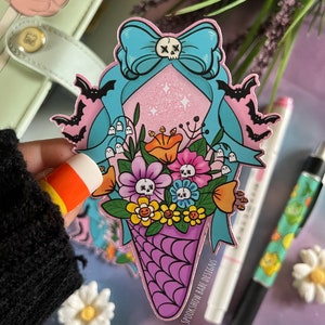 May include: A pink and blue sticker featuring a skull and crossbones bow, bats, flowers, and a purple and white ice cream cone. The sticker reads "SpookShow Babe Designs".