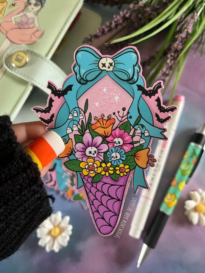 May include: A pink and blue sticker featuring a skull and crossbones bow, bats, flowers, and a purple and white ice cream cone. The sticker reads "SpookShow Babe Designs".