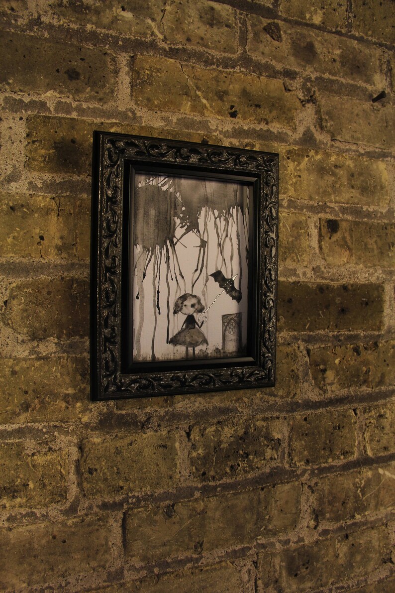 May include: A black framed picture with a white background and a black and white illustration of a girl holding a wand with a bat flying above her. The illustration is surrounded by black ink splatters.