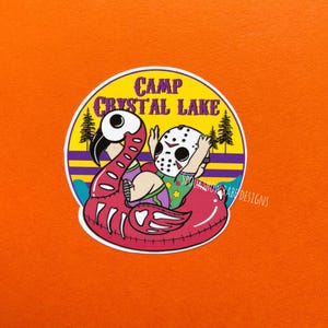 May include: A round sticker with a cartoon illustration of a character in a hockey mask floating on a pink flamingo pool float. The sticker has the text "Camp Crystal Lake" in a yellow and purple design.