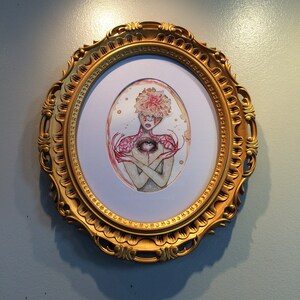 May include: A framed watercolour painting of a person with a large eye in the centre of their chest. The person is surrounded by pink flowers and has a crown of flowers on their head. The frame is gold and ornate.
