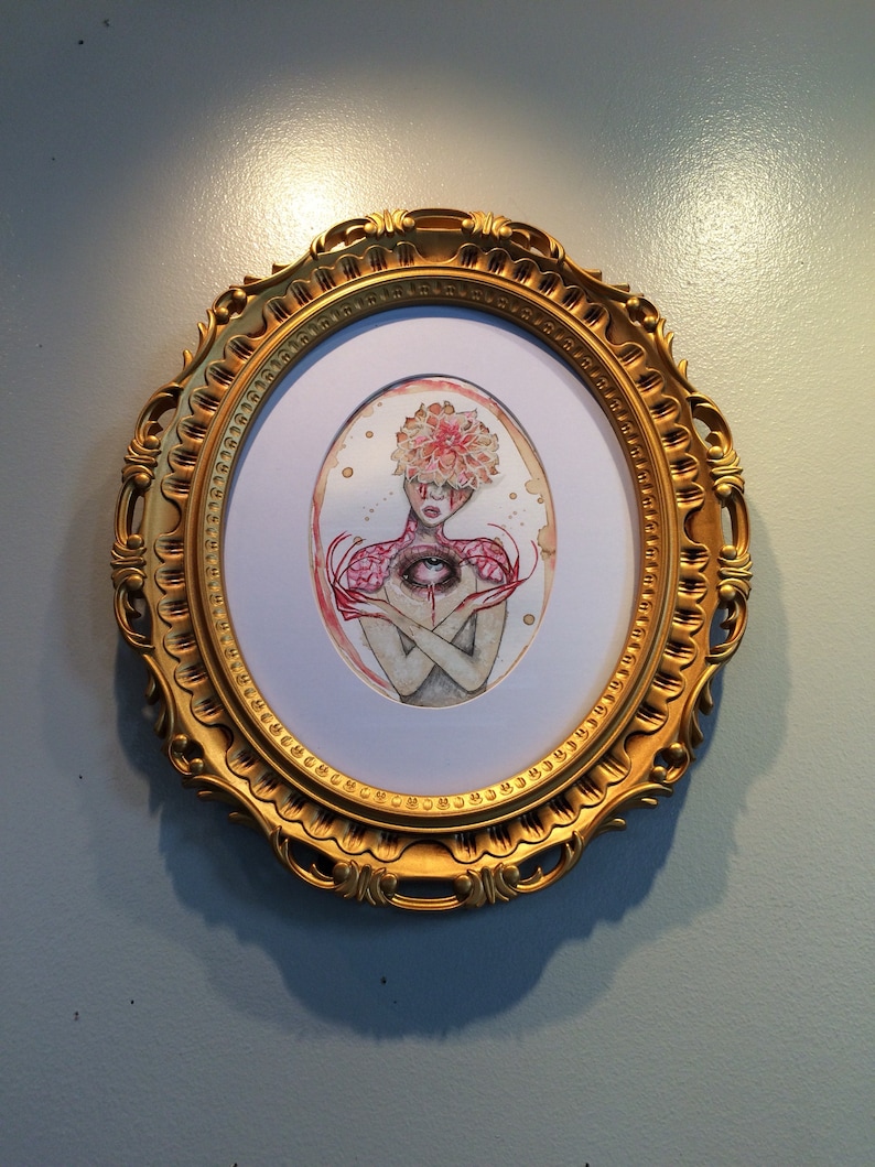 May include: A framed watercolour painting of a person with a large eye in the centre of their chest. The person is surrounded by pink flowers and has a crown of flowers on their head. The frame is gold and ornate.