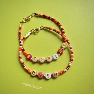 Puede incluir: Dos pulseras de cuentas con cierres dorados y cuentas de letras que deletrean "BONFIRE BASH". Las pulseras presentan cuentas naranjas, rosas, blancas y marrones. El fondo es de color verde claro liso.
