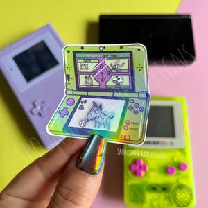 May include: A holographic sticker of a green Nintendo DS with a cartoon dog on the screen. The DS screen displays the date "2002 06:45" and the text "Nintendo Jack Skellington".
