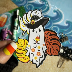 May include: A white ghost wearing a black witch hat with a spiderweb and a sunflower on top. The ghost is holding a candy corn popsicle and is surrounded by tropical leaves and bats. The text "SPOOKSHOW BABE DESIGNS" is in the top right corner of the image.