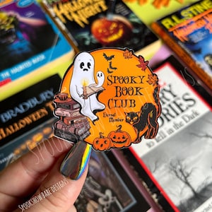 May include: A spooky book club enamel pin featuring a ghost reading a book, a black cat, and two jack-o'-lanterns. The pin is orange and black with the text "Spooky Book Club" and "Eternal Member".