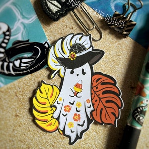 May include: A white ghost wearing a black witch hat with a spiderweb and a sunflower on top. The ghost is holding a popsicle and has a tropical leaf design on its body. The ghost is surrounded by tropical leaves.