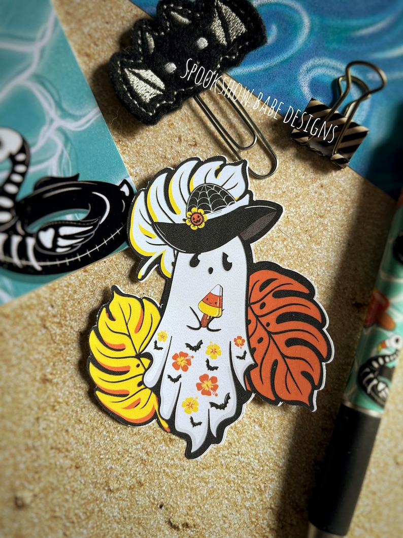 May include: A white ghost wearing a black witch hat with a spiderweb and a sunflower on top. The ghost is holding a popsicle and has a tropical leaf design on its body. The ghost is surrounded by tropical leaves.