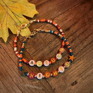 May include: Two beaded bracelets spelling "AUTUMN NIGHTS" with colourful letter beads. The bracelets have gold clasps and star charms. The background is a wooden surface with a fall leaf.