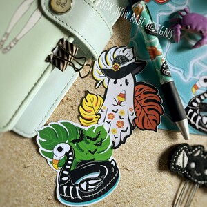 May include: A set of four Halloween-themed stickers featuring a ghost wearing a witch hat, a skeleton in a pool float, and two tropical leaves with bats. The stickers are on a light brown sandy background.