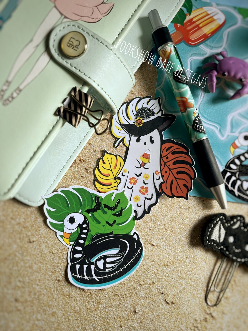 May include: A set of four Halloween-themed stickers featuring a ghost wearing a witch hat, a skeleton in a pool float, and two tropical leaves with bats. The stickers are on a light brown sandy background.