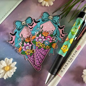 May include: Two pink and purple cone-shaped bouquets with a skull and crossbones design. The bouquets are decorated with blue bows, bats, and flowers. The bouquets are on a purple and blue background with white daisies.