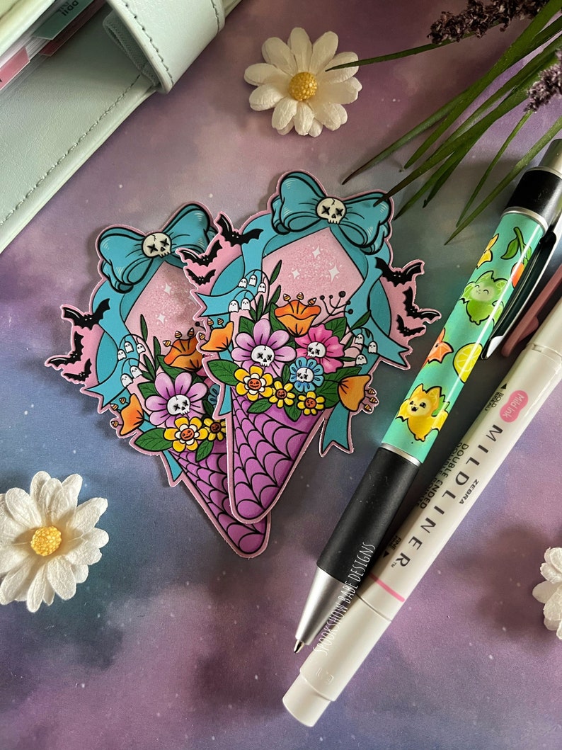 May include: Two pink and purple cone-shaped bouquets with a skull and crossbones design. The bouquets are decorated with blue bows, bats, and flowers. The bouquets are on a purple and blue background with white daisies.