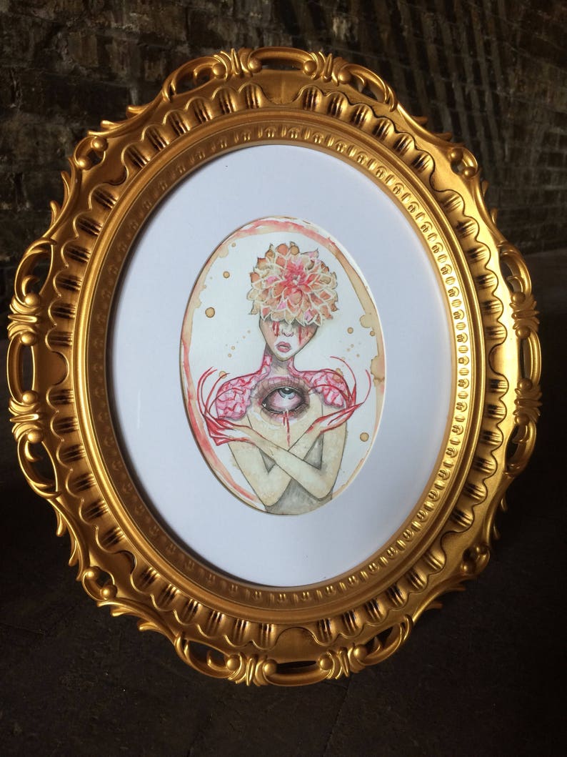 May include: A watercolour painting of a person with a flower head and an eye in their chest. The painting is framed in a gold oval frame.