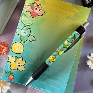 May include: A stationery set featuring a notepad and a matching pen. The notepad has a yellow-to-teal gradient with orange, lime, and lemon fruit illustrations, small bats, and orange horizontal lines. The text SPOOKSHOW BABE DESIGNS is at the bottom.