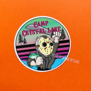 May include: A round sticker with a cartoon illustration of a character wearing a hockey mask and holding a machete. The sticker has the text "Camp Crystal Lake" and a small green frog. The background is orange.