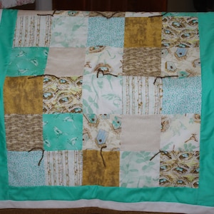 May include: A patchwork quilt with a variety of patterned squares in shades of teal, tan, and white. The quilt features bird and nest motifs, with a teal border and white trim. The quilt is approximately 100 cm by 100 cm.