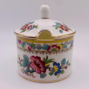Op de afbeelding: Een witte keramische suikerpot met deksel, versierd met kleurrijke bloempatronen. De pot heeft een gouden rand en een klein handvat op het deksel. Het bloemmotief omvat roze, blauwe en rode bloemen met groene bladeren.