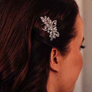 May include: A silver hair comb with a floral design featuring clear rhinestones. The comb is worn in a woman's dark brown hair.