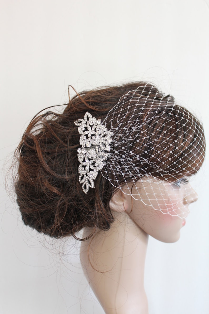 May include: A white bridal veil with a delicate net pattern, worn over a brown updo. A silver hair comb with crystal embellishments is pinned to the hair.
