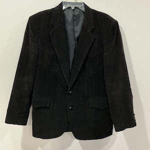 Puede incluir: Un blazer de pana negro con cierre de dos botones, solapas con muescas y dos bolsillos delanteros. El blazer tiene un diseño clásico con costuras decorativas en la parte delantera y los puños.