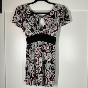 Puede incluir: Vestido de manga corta con estampado floral en negro, blanco y burdeos. El vestido presenta un escote en forma de ojo de cerradura y una banda negra en la cintura. El vestido está colgado de una percha negra sobre un fondo blanco.