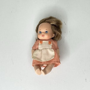 May include: A small doll with blue eyes and light brown hair. The doll wears a peach-coloured dress with a cream-coloured apron and orange buttons. The doll's arms and legs are slightly bent, and it is lying down.