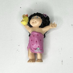 Puede incluir: Una figura de Cabbage Patch Kids de un niño envuelto en una toalla rosa con lunares blancos, sosteniendo un patito de goma amarillo. El niño tiene cabello negro rizado y un pequeño lazo rosa. La figura tiene una pequeña etiqueta.