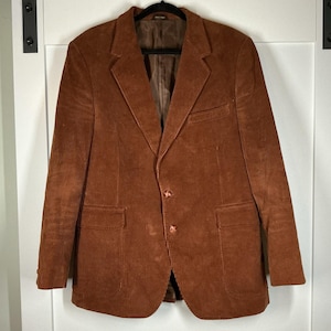 May include: A brown corduroy blazer with two buttons, two front pockets, and a notched lapel. The jacket is hanging on a black hanger against a white background. The blazer has a classic, tailored design.