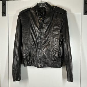 May include: A black leather jacket with a stand-up collar and shoulder epaulettes. The jacket features a button-up front, two front pockets, and a fitted shape. The leather has a worn appearance, suggesting a vintage style.