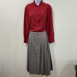 May include: A vintage outfit comprising a long-sleeved, red button-down shirt and a knee-length plaid skirt. The skirt has a grey, black, and red pattern. The shirt is buttoned up and has a high collar. The outfit is displayed on a mannequin.