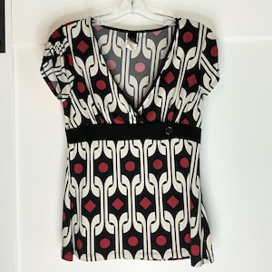 Puede incluir: Top de manga corta con cuello en V y un estampado geométrico en negro, blanco y rojo. El top tiene una banda negra en la cintura con un solo botón. El estampado incluye círculos y diamantes rojos, y líneas curvas blancas y negras.