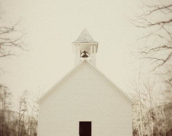 May include: A white wooden church with a bell tower and a single door. The church is surrounded by trees and the sky is a light gray.