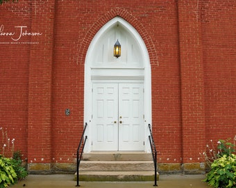 May include: A white double door with a small window above it, set in a red brick wall. The door is framed by a white archway and has a black metal railing on either side. The number 16 is visible on the brick wall.