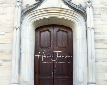 May include: A pair of wooden double doors with a decorative archway above them. The doors are brown and have a simple design. The archway is made of stone and has a cross at the top. The doors are set in a stone wall.