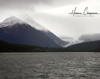 May include: A view of a glacier nestled between two snow-capped mountains. The glacier is visible in the distance, with a body of water in the foreground.