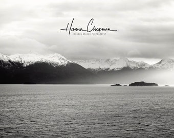 May include: A black and white photograph of a snow-capped mountain range in the distance, with a small island in the foreground. The water is calm and the sky is overcast. The text "Hanna Chapman, Johnson Branch Photography" is in the upper center of the image.
