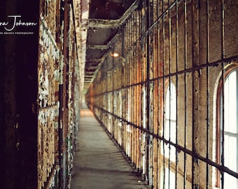 May include: A long, narrow hallway in an abandoned prison with barred windows and peeling paint on the walls. The hallway is empty and the light is dim.