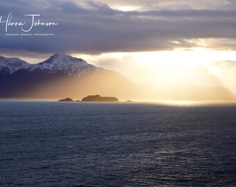May include: A panoramic view of a mountain range with snow-capped peaks, a small island in the foreground, and a bright, golden sun shining through the clouds over a calm, blue ocean.