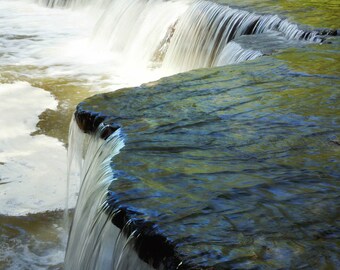 May include: A cascading waterfall flows over a series of rock formations, creating a picturesque scene. The water is clear and sparkling, and the rocks are covered in moss and lichen. The image is taken from a low angle, giving the viewer a sense of the waterfall's power and beauty.