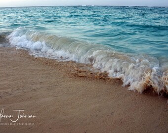 May include: A wave crashes on a sandy beach, creating a foamy white crest against the turquoise water. The sand is a light brown color, and the water is clear and inviting.