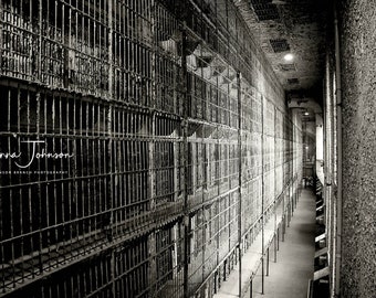 May include: Black and white photo of a long hallway with rows of barred cells on one side and a rough brick wall on the other. The hallway is lit by a single lightbulb at the end. The text "Hanna Johnson" and "Johnson Branch Photography" is visible in the lower left corner.