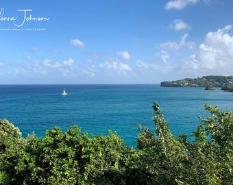 May include: A sailboat on a calm blue ocean with a lush green island in the distance. The sky is a bright blue with white clouds.