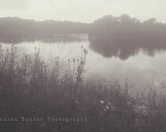 May include: A black and white photograph of a lake with a small island in the middle. The lake is surrounded by trees and the water is calm. The photo is taken from the shore of the lake, looking out towards the island. The photo is taken in a foggy or misty environment.