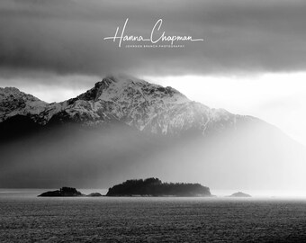 May include: A black and white photograph of a snow-capped mountain range with a small island in the foreground. The mountain range is partially obscured by clouds, and the island is surrounded by water. The image is taken from a distance, and the mountains are in the background.